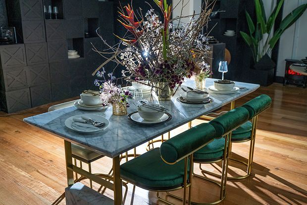 A styled dining room featuring a long rectangular marble dining table with a gold-toned metal base, set with layered plates, bowls, glassware and cutlery. Deep emerald green upholstered dining chairs with rounded backs surround the table. A large dramatic floral arrangement forms the centrepiece, while dark built-in wall cabinetry and warm timber flooring create a sophisticated, richly contrasted interior.