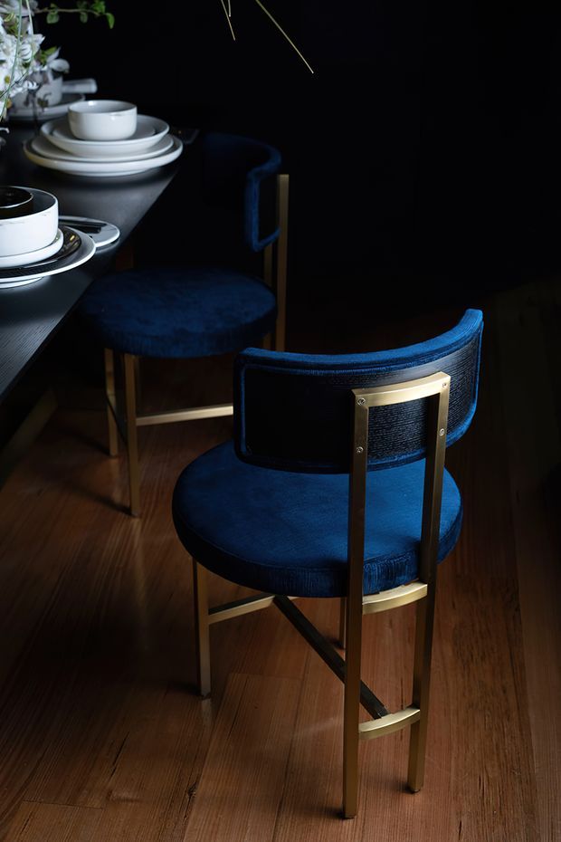 A moody interior detail showing two deep navy upholstered dining chairs with dark wood and brass-toned metal frames beside a dark dining table. White ceramic plates, bowls, and cups are arranged on the tabletop, with warm timber flooring below and strong directional lighting against a nearly black background.