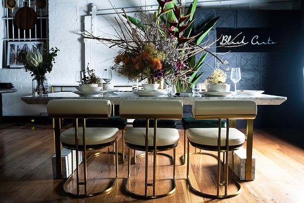 A stylish dining area with a long stone-topped dining table and three upholstered bar-height chairs with brass-toned metal frames. The room features dark timber flooring, white painted brick walls, a dark geometric feature wall with cursive lettering, open shelving and abundant sculptural floral arrangements. Strong natural light creates a refined, high-contrast atmosphere.