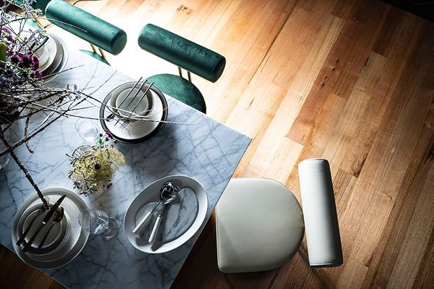 Overhead view of a refined dining area with a rectangular grey-white veined stone dining table, sculptural green upholstered dining chairs, and a light upholstered chair at the table end. Place settings, glassware, and organic branch arrangements sit on the table, while warm timber flooring surrounds the furniture.