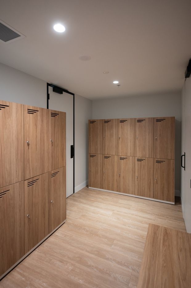 A clean, functional locker room with rows of light timber-look lockers featuring black ventilation slots and small metal handles. The room has pale wood-look flooring, white walls and ceiling, recessed downlights, a wall-mounted air vent, and a simple white door with black hardware.