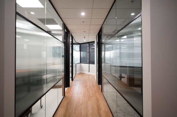 A modern commercial office corridor lined with black-framed glass-partitioned workspaces. The hallway has warm wood-look flooring, white acoustic ceiling tiles with recessed lighting, and translucent privacy film on the glazing. Reflections reveal additional office desks and rooms.