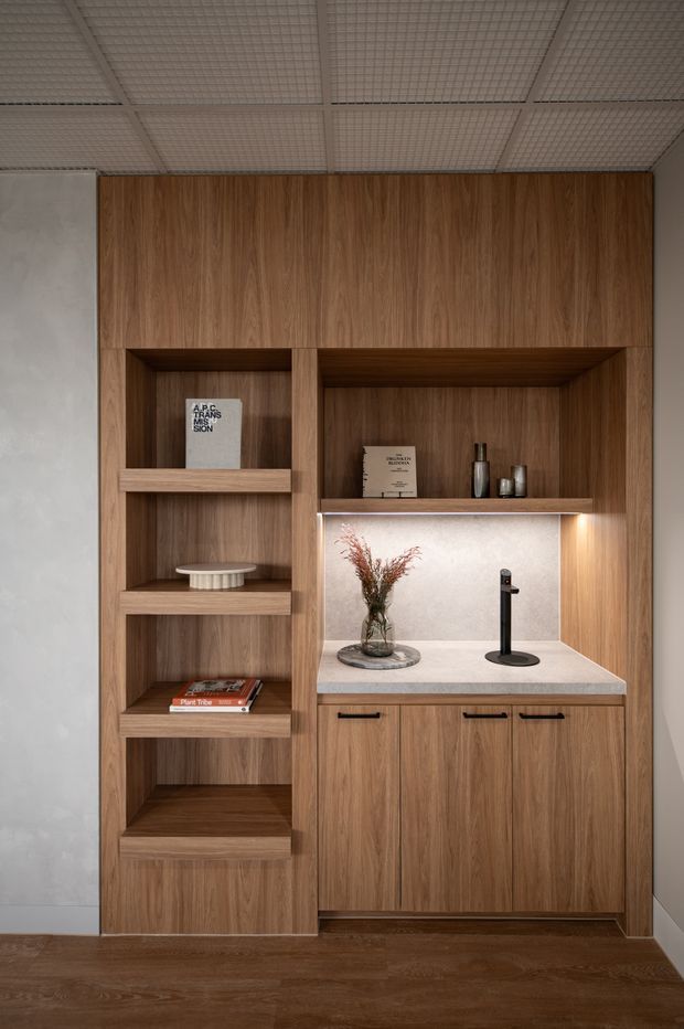 A compact built-in timber joinery unit combines open display shelving with a recessed beverage station and closed base cabinets. The warm wood grain cabinetry frames a pale stone benchtop and illuminated backsplash, with a black mixer tap, small decorative vessels, books, and a vase. The space has a grid-pattern suspended ceiling, dark timber flooring, and a restrained contemporary fit-out.