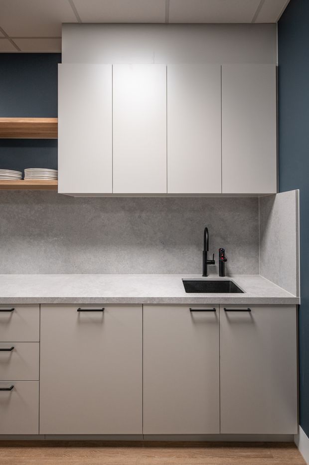 Compact contemporary kitchenette with matte light-grey flat-panel cabinetry, a pale stone-look benchtop and matching grey splashback. A black undermount sink and tap provide contrast, while open timber shelving with stacked plates is visible at the left. The space has dark blue-grey walls, a suspended grid ceiling and timber-look flooring.