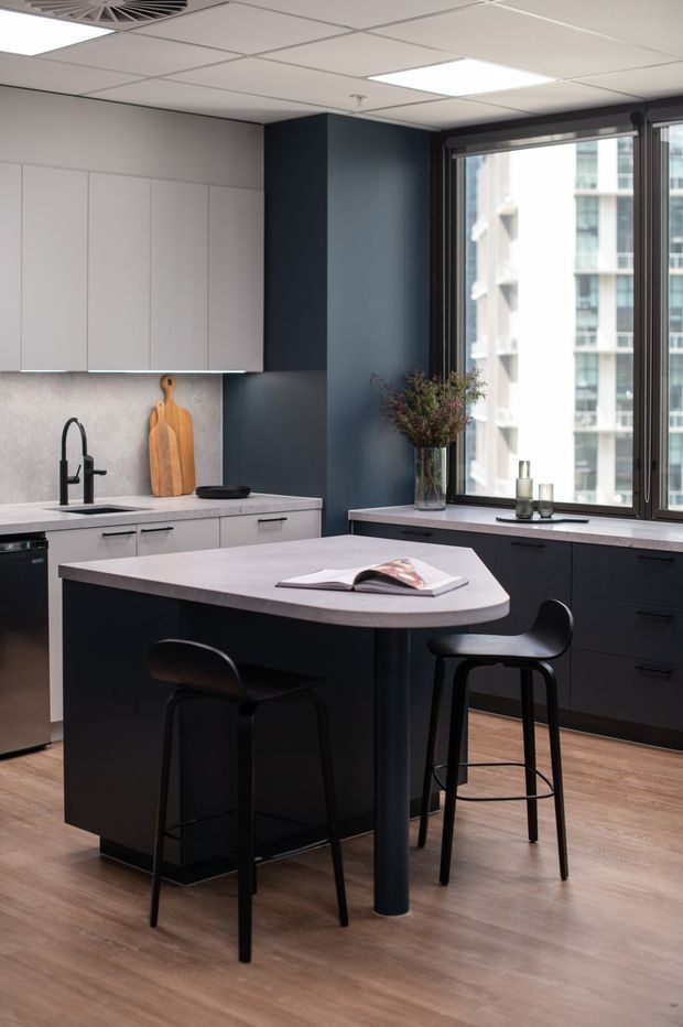 A compact contemporary kitchen in an urban apartment, featuring a dark navy island and lower cabinetry with pale grey-white upper cabinets and benchtops. The island has a rounded end and seating for two at black bar stools. A black tap, timber chopping boards, integrated appliances, and a large window overlooking neighbouring high-rise buildings complete the space. The room has a suspended white grid ceiling with recessed panel lighting and warm timber-look flooring.