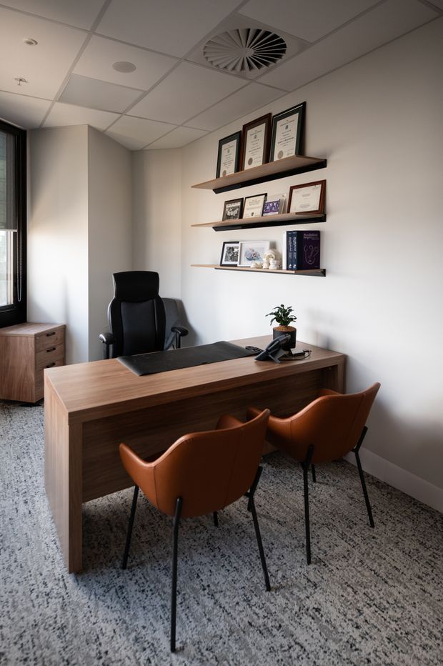 A compact, professionally furnished private office with a large dark timber desk, two visitor chairs, and a black executive chair. Floating timber shelves display framed certificates and books on a light neutral wall. The room has speckled carpet, a suspended acoustic ceiling with integrated HVAC, a narrow window, and understated contemporary detailing.