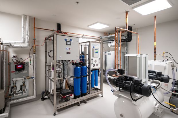 Bright, utilitarian plant room filled with exposed copper and metal pipework, filtration vessels, control panels, pumps, pressure equipment and mechanical services. The room has smooth white walls and flooring, recessed square ceiling lights, and a highly technical commercial or industrial fit-out.