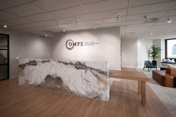A contemporary commercial reception and waiting area with a large marble reception desk, timber-look flooring, timber meeting table, neutral plastered walls, and a suspended acoustic tile ceiling. Seating, a plant, and city views are visible in the adjacent lounge area.