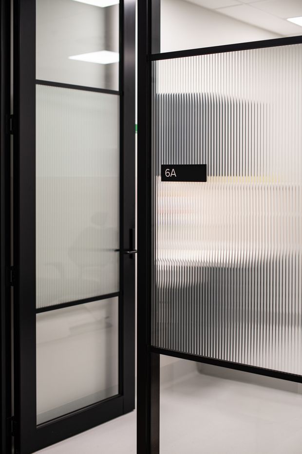 Close-up view of a contemporary commercial office entry or partition system with black metal-framed doors and ribbed translucent privacy glazing. A black sign marked “6A” is mounted on the glass, with a bright, minimal office interior visible through the textured glazing.
