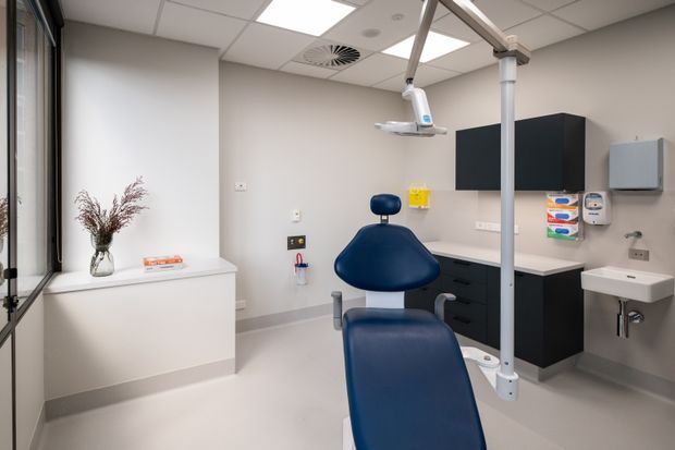 A clean, compact clinical treatment room, likely a dental or medical surgery, viewed from the entry. The room contains a dark blue adjustable treatment chair, overhead examination light, wall-mounted cabinetry and workbench, hand basin, clinical dispensers, and a suspended acoustic tile ceiling. Neutral grey and white finishes create a bright, functional healthcare environment.