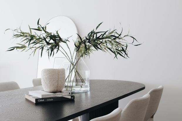 A refined dining area featuring a dark rectangular dining table with a subtly textured surface, upholstered light beige dining chairs, and a clear glass vase with leafy branches as a centrepiece. Minimal white walls and a large round mirror create a calm, understated setting, with stacked design books adding a small styling detail.