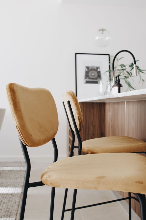 A bright, minimalist dining area featuring tan upholstered bar stools with black metal frames positioned at a wood-fronted kitchen island or breakfast bar. The island has a white stone-look benchtop, while a framed vintage camera print, curved black-framed mirror, pendant light, and potted greenery decorate the white wall behind.
