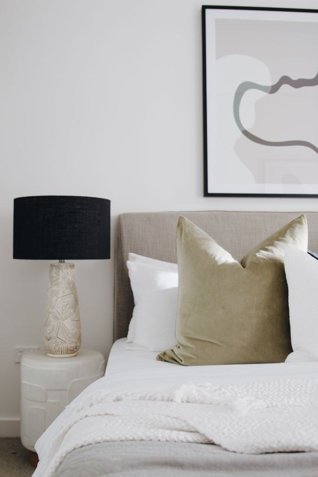 A softly styled bedroom corner featuring a neutral upholstered bedhead, layered white bedding, a large taupe accent cushion, and a white sculptural bedside table with a patterned ceramic lamp and black drum shade. Minimal abstract artwork in a black frame hangs above the bed, creating a calm, refined interior.