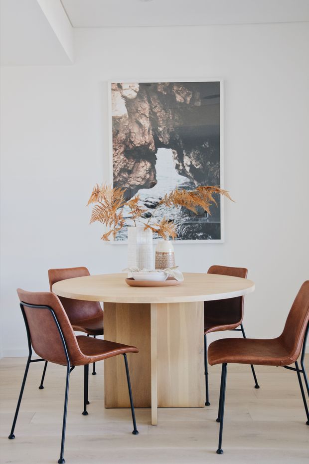 Bright, minimalist dining area with a round light-wood pedestal table surrounded by brown leather dining chairs with slim black metal legs. A large framed nature photograph hangs on the white wall, while dried foliage and neutral ceramics form a simple tabletop arrangement. Pale timber flooring and white walls create an airy, calm setting.