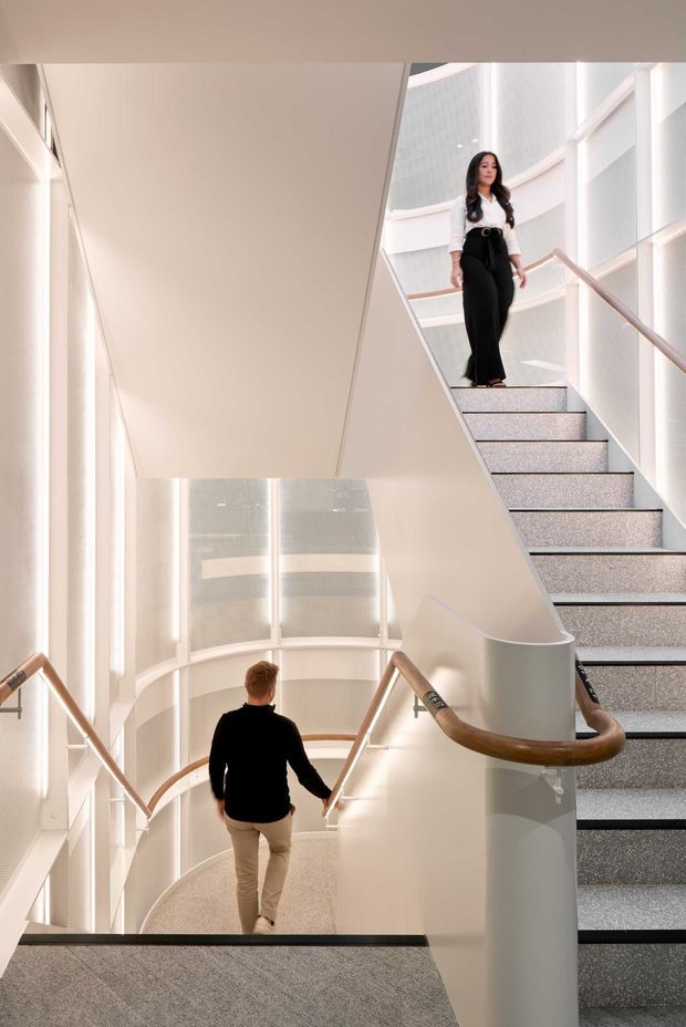 A bright, multi-level interior stairway with intersecting stair flights, pale curved walls, tall vertical glazing, and warm timber handrails. Grey carpeted steps and landings contrast with the predominantly white architectural surfaces. Two people are shown using the stairs, emphasizing the circulation and generous vertical space.