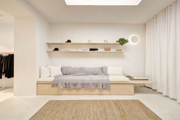 Bright minimalist living room with a built-in pale timber daybed or sofa beneath two floating display shelves. The room has white walls, full-height sheer curtains, a large skylight or ceiling light, pale flooring, sparse decor, and a woven rectangular rug. A walk-in wardrobe or clothing area is visible through the opening on the left.
