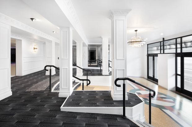A bright, refined interior corridor or lobby with white decorative wall panelling, ornate cornices, classical pilasters and columns, black handrails, and short level changes connected by steps and ramps. Dark patterned carpet contrasts with light terrazzo flooring, while black-framed glazed partitions and doors define a contemporary area on the right. A crystal chandelier and wall sconces add an elegant decorative character.