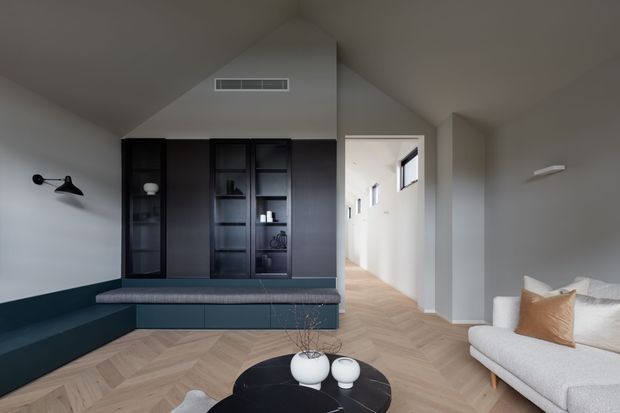 Contemporary living room with a vaulted ceiling, pale grey walls, dark built-in cabinetry and integrated bench seating, a light upholstered sofa, black round coffee tables, and warm herringbone timber flooring. A bright hallway extends beyond the room.