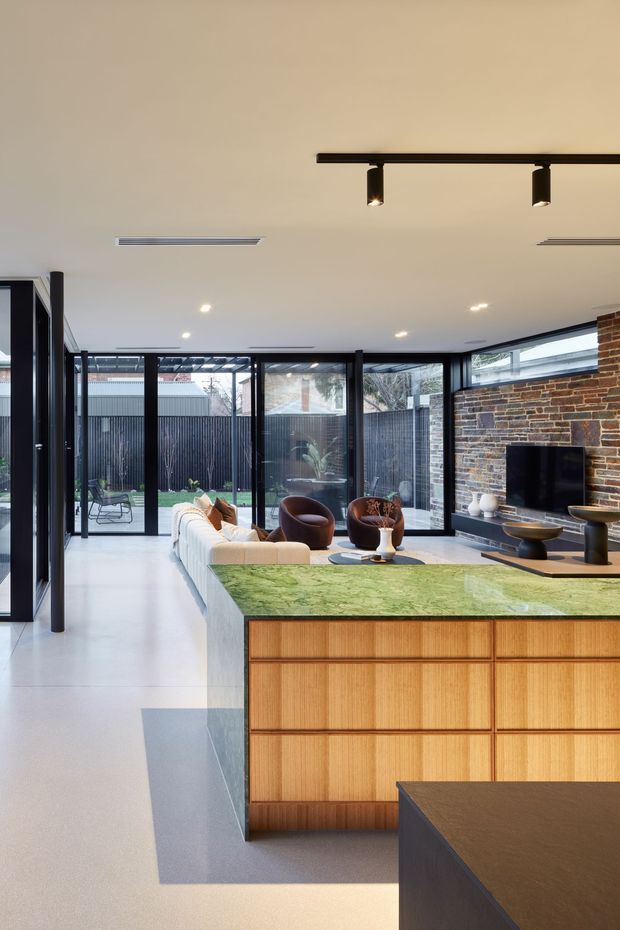 Contemporary open-plan kitchen and living area with a large green stone island, warm timber cabinetry, pale polished flooring, black-framed floor-to-ceiling glazing, and an exposed brick feature wall with a built-in fireplace or television. The glazing opens toward a small landscaped courtyard, while sculptural lounge furniture and track lighting complete the refined interior.