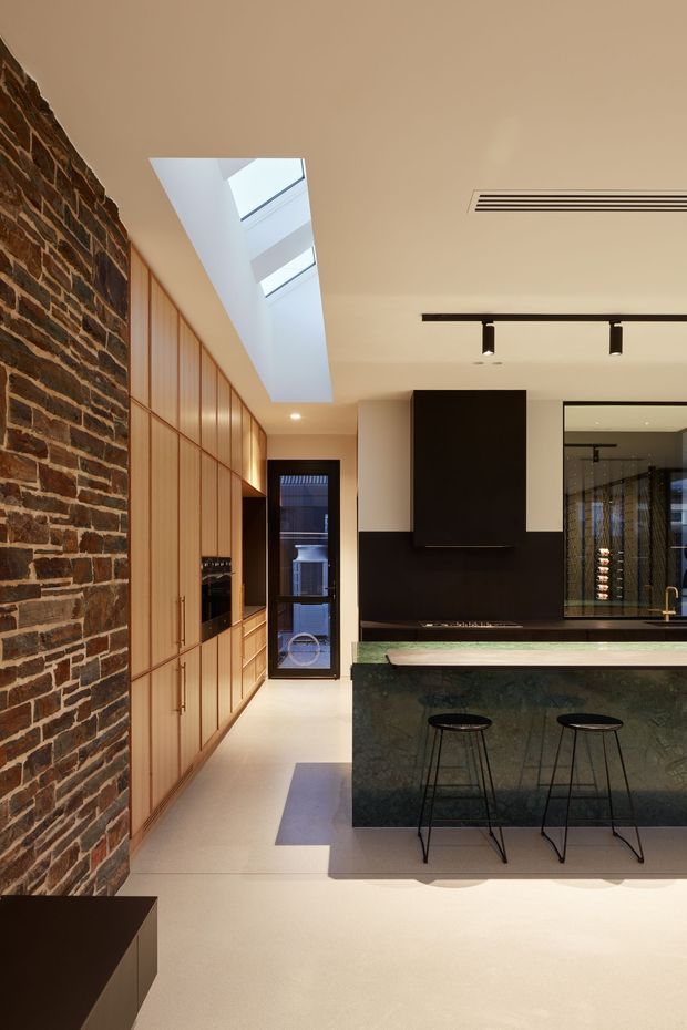 A refined contemporary kitchen with a long timber-lined cabinetry wall, exposed dark brick feature wall, and a large dark green stone island with integrated seating. Black cabinetry, fittings, track lighting, and a linear air-conditioning vent create a strong modern contrast, while a skylight brings natural light into the space. A glimpse of a wine display or beverage area is visible through the glazing at the rear.