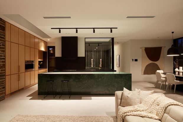 Open-plan contemporary kitchen and living area with a large dark green stone island, extensive warm timber cabinetry, integrated appliances, black pendant and track lighting, and a partially visible dining and lounge zone. A glazed wine display or cellar feature forms a focal point behind the island, while neutral furnishings and abstract wall art create a refined, warm interior.