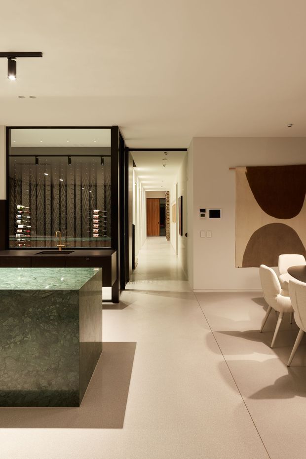 A refined open-plan interior combining a kitchen, dining area and hallway. The kitchen features a large green stone island with an integrated sink and brass-toned tap, dark cabinetry, and black-framed glazed wine storage. Pale smooth flooring and warm off-white walls create a bright, softly minimalist setting. A sculptural abstract artwork and curved upholstered dining chairs add understated decorative detail.