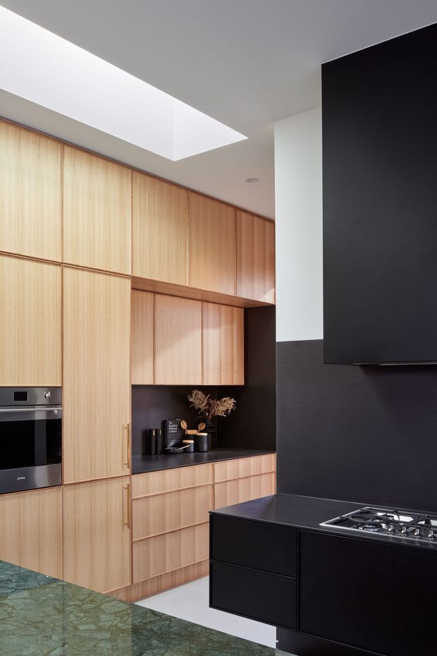 A contemporary kitchen with full-height light timber cabinetry, integrated black appliances, a dark stone or composite benchtop, and a black kitchen island with gas cooktop. A large rectangular skylight brings natural light into the space, contrasting with the dark feature wall and cabinetry accents. A green-toned stone surface is visible in the foreground.