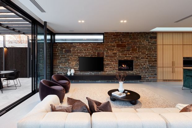 A refined contemporary living room with a large cream sectional sofa and sculptural dark lounge chairs arranged around a low black coffee table. The room features a full-height exposed brick feature wall with a wall-mounted television and built-in fireplace, extensive black-framed glazing opening to an outdoor area, pale flooring, and warm timber cabinetry at the right. A compact kitchen zone is partially visible beyond the living area.