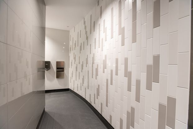 A narrow, modern interior corridor with a grey floor, dark skirting, and predominantly white tiled walls. The right wall features a textured geometric arrangement of raised and recessed rectangular tiles in white and light grey, while the left wall has large smooth pale tiles. Minimal wall-mounted fixtures are visible at the far end, with recessed ceiling lighting creating an even, clean appearance.