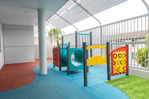 A bright, covered children's play area with colourful fixed playground equipment, including a crawl tunnel, activity panels and climbing elements. The space has a blue and red safety surface, a small artificial grass patch, white vertical railings, a gridded overhead canopy, and a white structural column and walls.