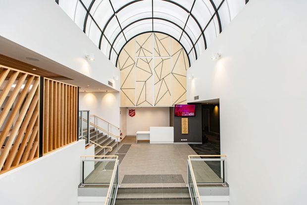 A bright, contemporary multi-level interior entrance or reception space with a dramatic barrel-vaulted glazed roof, white walls, grey tiled flooring, and a large geometric timber feature panel. Stairs descend in the foreground beside glass balustrades, while a timber slatted screen, reception counter, digital display, and dark doorway define the lobby area.