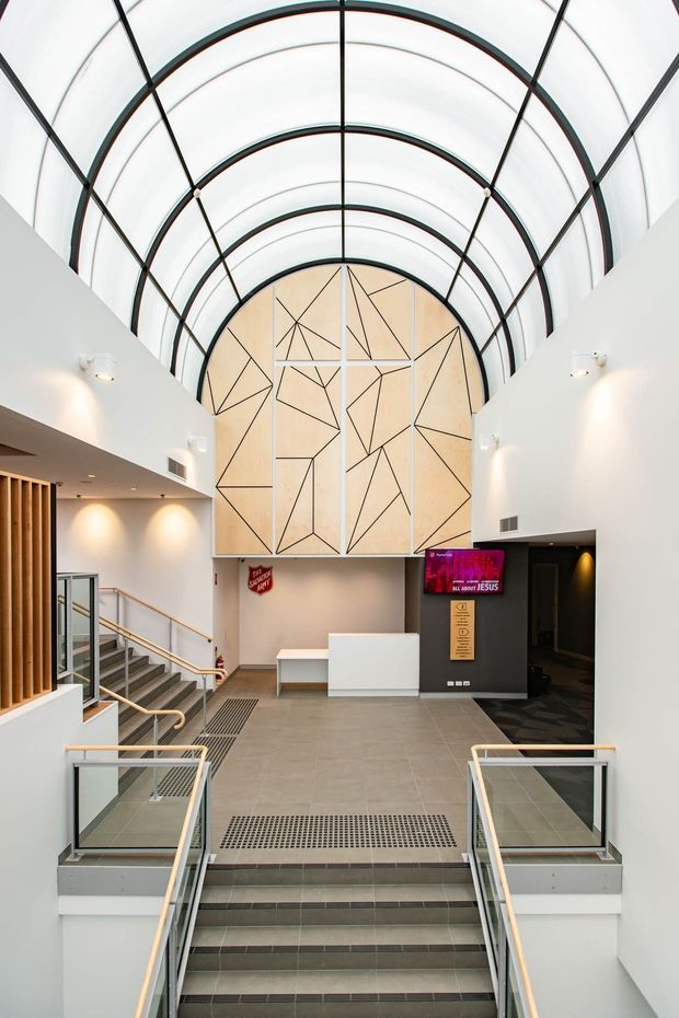 Bright contemporary multi-level commercial interior, likely a public foyer or entry hall, viewed from a stair landing. The space features a dramatic barrel-vaulted translucent ceiling with black framing, white walls, grey tiled floors, glass and pale timber balustrades, a reception counter, a black doorway surround, and a large geometric timber-panel feature wall. Stairs and accessible circulation routes dominate the foreground.