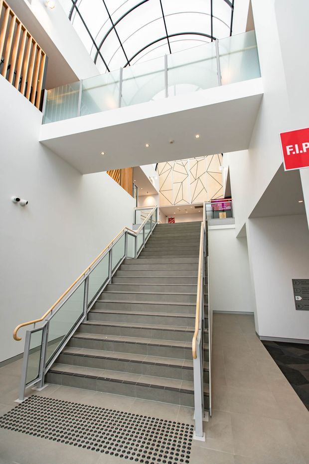 A bright, contemporary multi-level interior stairway in a large public or commercial building. The broad tiled staircase has glass balustrades, pale timber handrails, and integrated accessibility details. White walls, an overhead bridge, a frosted-glass mezzanine balustrade, and a dramatic curved glazed roof create a tall atrium-like circulation space. A geometric feature wall and directional signage are visible at the upper landing.