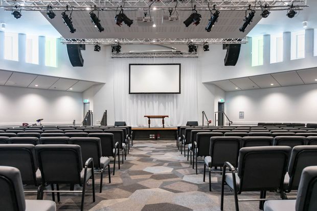 A bright, contemporary multipurpose auditorium or church hall viewed from the rear, with rows of black upholstered chairs facing a stage, projection screen, lectern/table, and extensive overhead theatrical lighting and speakers. The room has white walls and ceiling panels, tall narrow windows, and a geometric patterned carpet aisle.