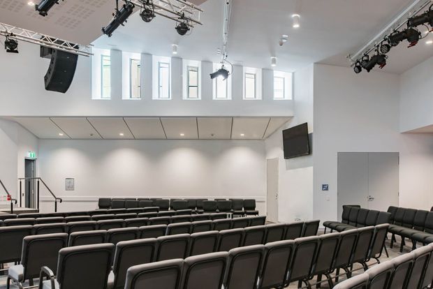 Bright modern auditorium or lecture theatre with rows of black upholstered seating, white walls, high ceiling, clerestory windows, a raised rear balcony, wall-mounted screens, and extensive stage or event lighting and speaker equipment.