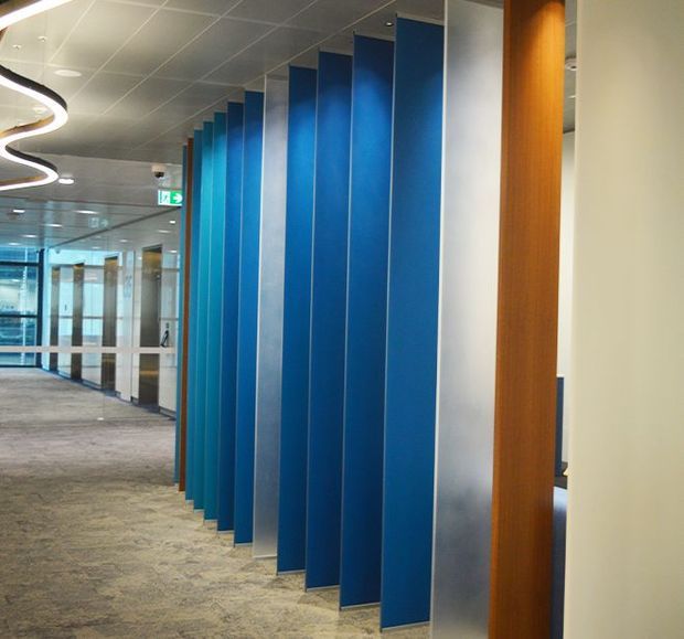 Modern commercial office corridor with a long sequence of vivid blue vertical partition fins, frosted or translucent panels, timber-framed openings, glass meeting-room fronts, carpet flooring, and a suspended grid ceiling. Curved contemporary ceiling lighting and strong linear perspective create a polished workplace interior.
