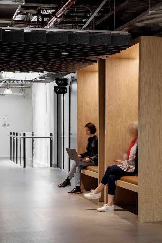 A contemporary commercial interior with an open-plan corridor and informal work or waiting booths. Full-height pale timber privacy panels form built-in seating niches, while dark slatted ceiling elements and exposed mechanical services create an industrial character. Two people are seated with a laptop, and a glazed balustrade overlooks a lower level or open area.