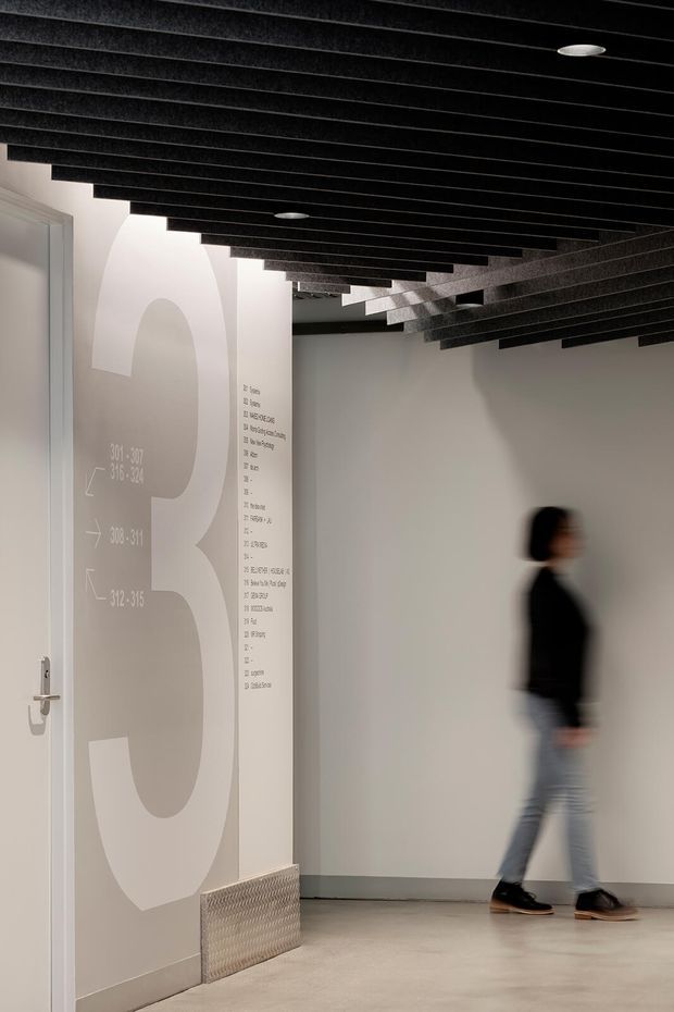 A modern interior corridor or commercial circulation space with pale grey walls, oversized wayfinding graphics and room numbers, a white door, polished concrete floor, and a dramatic black ceiling composed of closely spaced acoustic fins with recessed circular lights. A blurred person is walking through the hallway, emphasizing the active public or workplace setting.