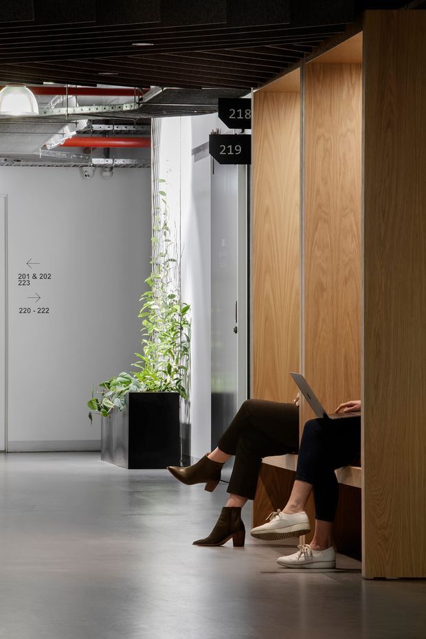 A contemporary commercial interior corridor or shared workspace with polished concrete flooring, exposed services and red ductwork, a dark linear slatted ceiling, grey wayfinding walls, and warm timber-panelled built-in seating or work booths. Two people are seated using laptops, with tall indoor plants adding greenery. Room numbers and directional signage indicate an office or institutional environment.