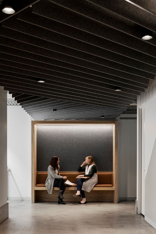 A contemporary commercial interior corridor or waiting area with a built-in timber-framed banquette and upholstered seating niche. Two people are seated facing each other beneath a dramatic exposed dark acoustic-batten ceiling with recessed and surface-mounted downlights. The surrounding walls are white, the floor is polished concrete, and the niche has a dark textured acoustic backdrop with warm timber and brown upholstery accents.