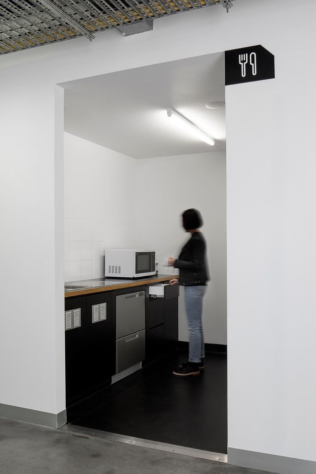 A compact commercial kitchenette or staff kitchen viewed through a wide doorway. The space has black built-in cabinetry and appliances, a timber-toned benchtop, white tiled and painted walls, black flooring, and exposed building services visible in the ceiling outside the room. A black sign with fork and knife icons identifies the area as a food-preparation space. A person is standing at the counter.