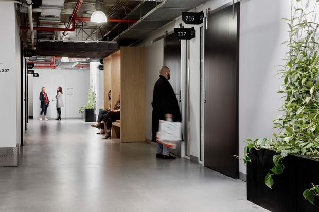 A modern commercial interior corridor with polished concrete flooring, dark metal-framed doors, light grey walls, exposed services and ceiling infrastructure, and black directional room-number signs. People are walking and waiting along the hallway, while large black planters with green foliage add contrast. The space has an industrial office or institutional character with a long, open circulation route.
