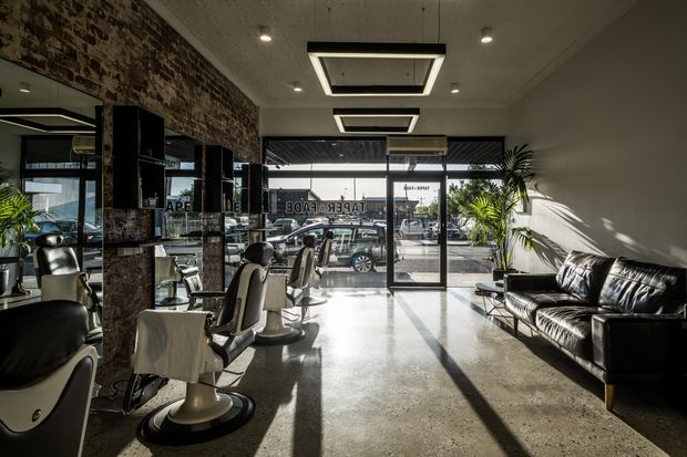 A bright contemporary commercial salon interior, likely a barber shop or hair salon, viewed toward a fully glazed street frontage. Multiple styling stations and salon chairs line the exposed brick wall on the left, while a black leather sofa sits against the white wall on the right. The polished concrete floor, exposed brick feature wall, black-framed glazing, and recessed geometric ceiling lights create an industrial-modern atmosphere. Strong sunlight enters through the storefront, casting long shadows across the floor.