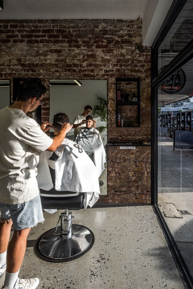 Interior view of a compact barber shop with exposed, weathered brick walls, large wall mirrors, recessed shelving, barber chairs and a concrete-look floor. A barber is cutting a client's hair, while strong sunlight enters through the full-height glazed storefront. The street and café signage are visible through the glass.