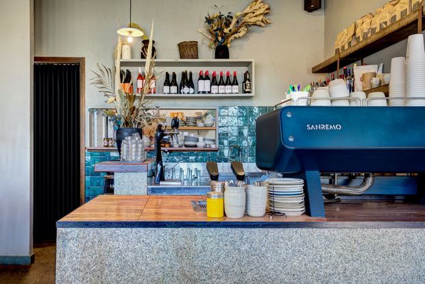 A compact, stylish café or coffee bar interior viewed from the customer side. A large blue espresso machine sits on a polished timber-topped counter with a speckled stone or terrazzo-style front. Behind it are turquoise glazed tile splashbacks, open shelving with cups, plates, bottles and decorative dried botanicals, creating a warm eclectic hospitality setting.