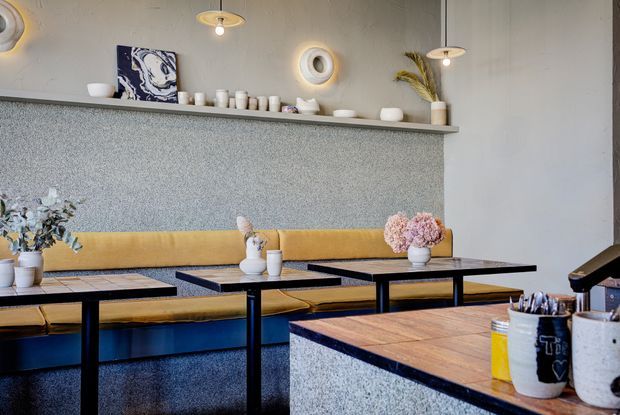 A stylish contemporary café or hospitality interior with a long mustard-yellow upholstered banquette, black-framed tables with timber and dark stone-like tops, and a textured grey service counter in the foreground. Open shelving displays ceramics and decorative objects, while sculptural pendant and wall lighting adds warmth. The palette combines mustard yellow, blue, grey, black, and natural timber accents.