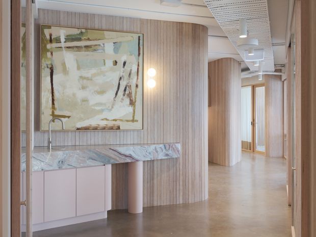 A refined contemporary interior hallway or entry zone with extensive vertical timber wall panelling, a compact built-in stone-topped kitchenette or bar cabinet, and a large abstract artwork. The space extends into a softly lit corridor with curved timber-clad walls, glazed doors, polished concrete flooring, and an exposed services ceiling with linear perforated panels and pendant lighting. The palette is warm and neutral with pale pink cabinetry, natural timber, grey stone, and concrete.