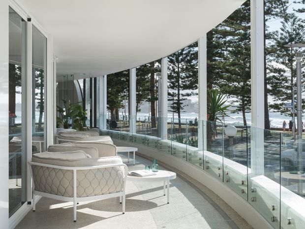 A bright, curved glazed terrace or sunroom overlooking a coastal setting. The space features floor-to-ceiling glass, white architectural finishes, a glass balustrade, pale upholstered lounge chairs, and a small white coffee table. Tall coastal trees and ocean views are visible beyond the glazing.