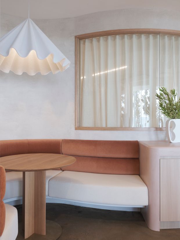 A softly styled built-in banquette dining nook with a curved terracotta leather or upholstered bench, pale upholstered seat cushions, a round timber pedestal table, and a pale timber-toned cabinet. The space has a white textured plaster wall, a large curved timber-framed window with sheer curtains, and a sculptural scalloped pendant light. The palette is warm, muted, and organic with contemporary rounded forms.