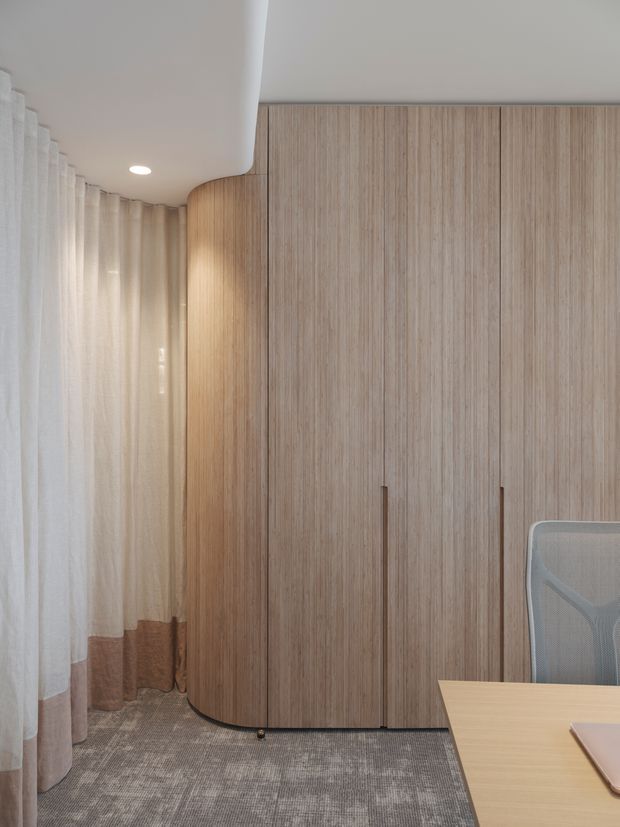 A refined, compact workspace with floor-to-ceiling light timber cabinetry featuring a curved corner, integrated vertical handles, sheer full-height curtains, a soft grey patterned carpet, and a light timber desk with a mesh office chair. The space has a calm, warm, minimalist contemporary character with recessed ceiling lighting.